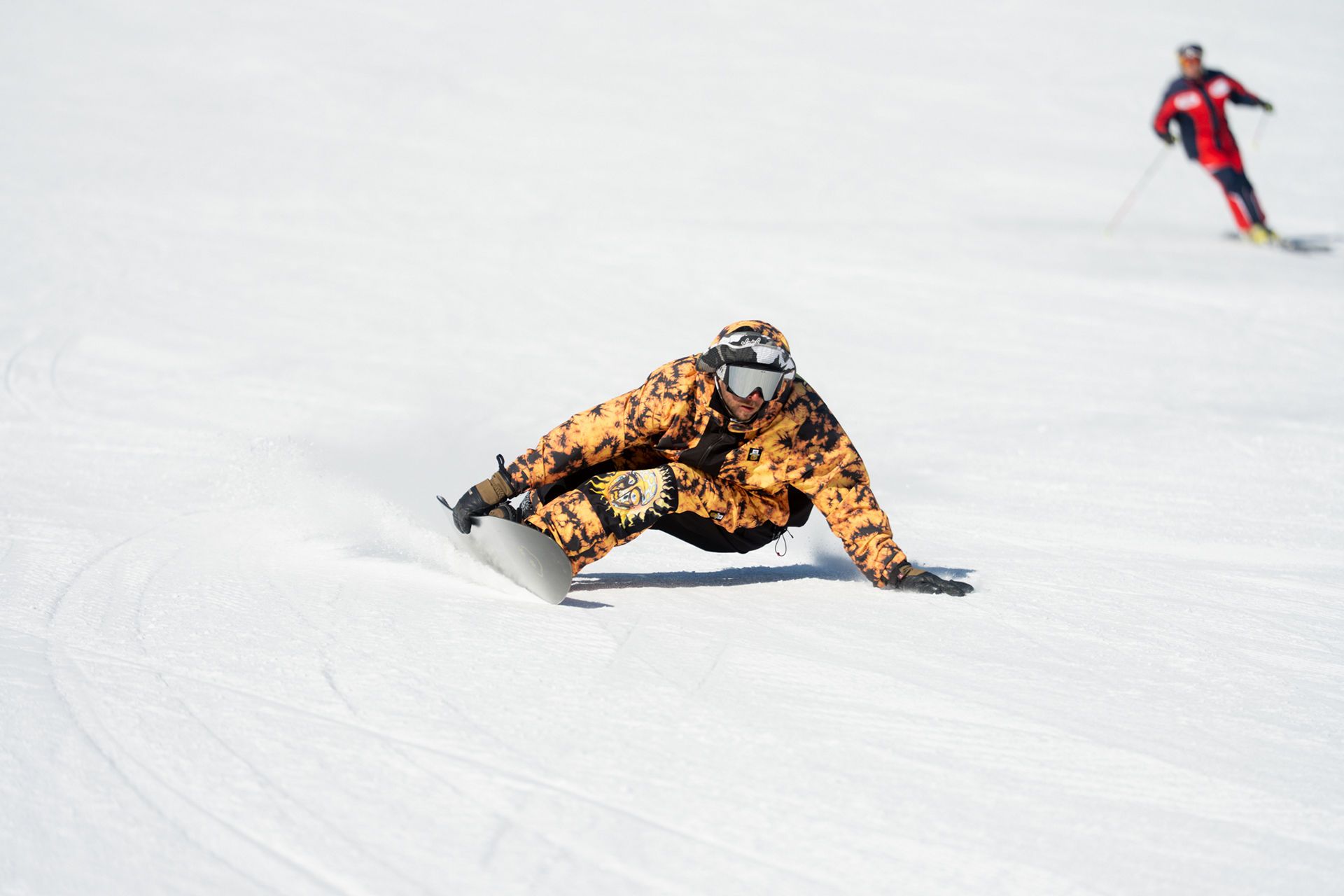 BLJ04778 snow vars | Fresh Planet | Fresh Planet Snowboarder in an orange camo jacket carving a turn on a snowy slope, wearing goggles and a helmet, with a skier in red in the background.