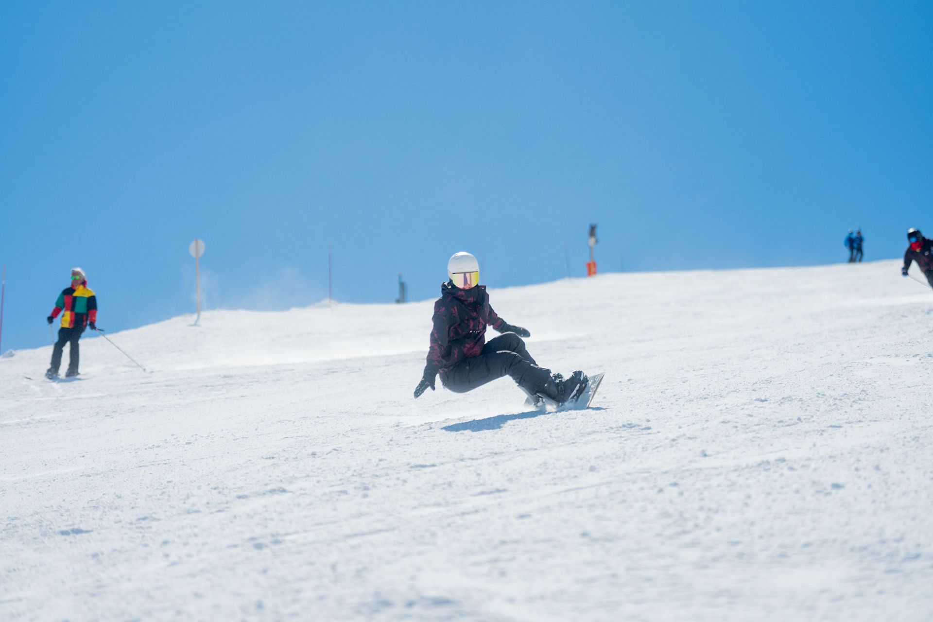 BLJ05693 snow vars | Fresh Planet | Fresh Planet Snowboarder in a dark jacket and white helmet carving down a snowy slope, with skiers in colorful outfits in the distance under a bright blue sky.