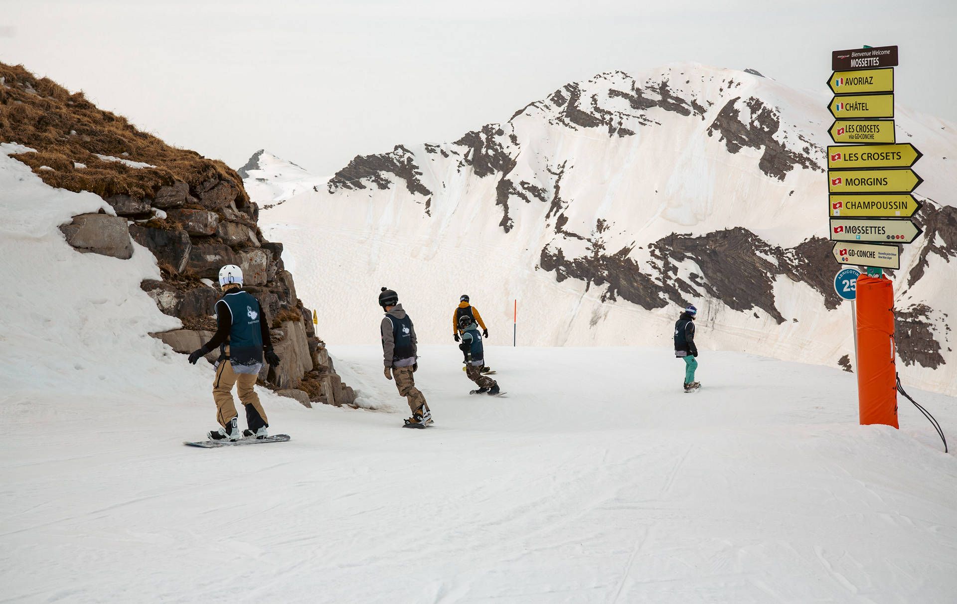FJ7B6124 snow avoriaz | Fresh Planet | Fresh Planet Group of snowboarders on a snowy mountain pass, with a tall signpost and yellow trail signs on the right.