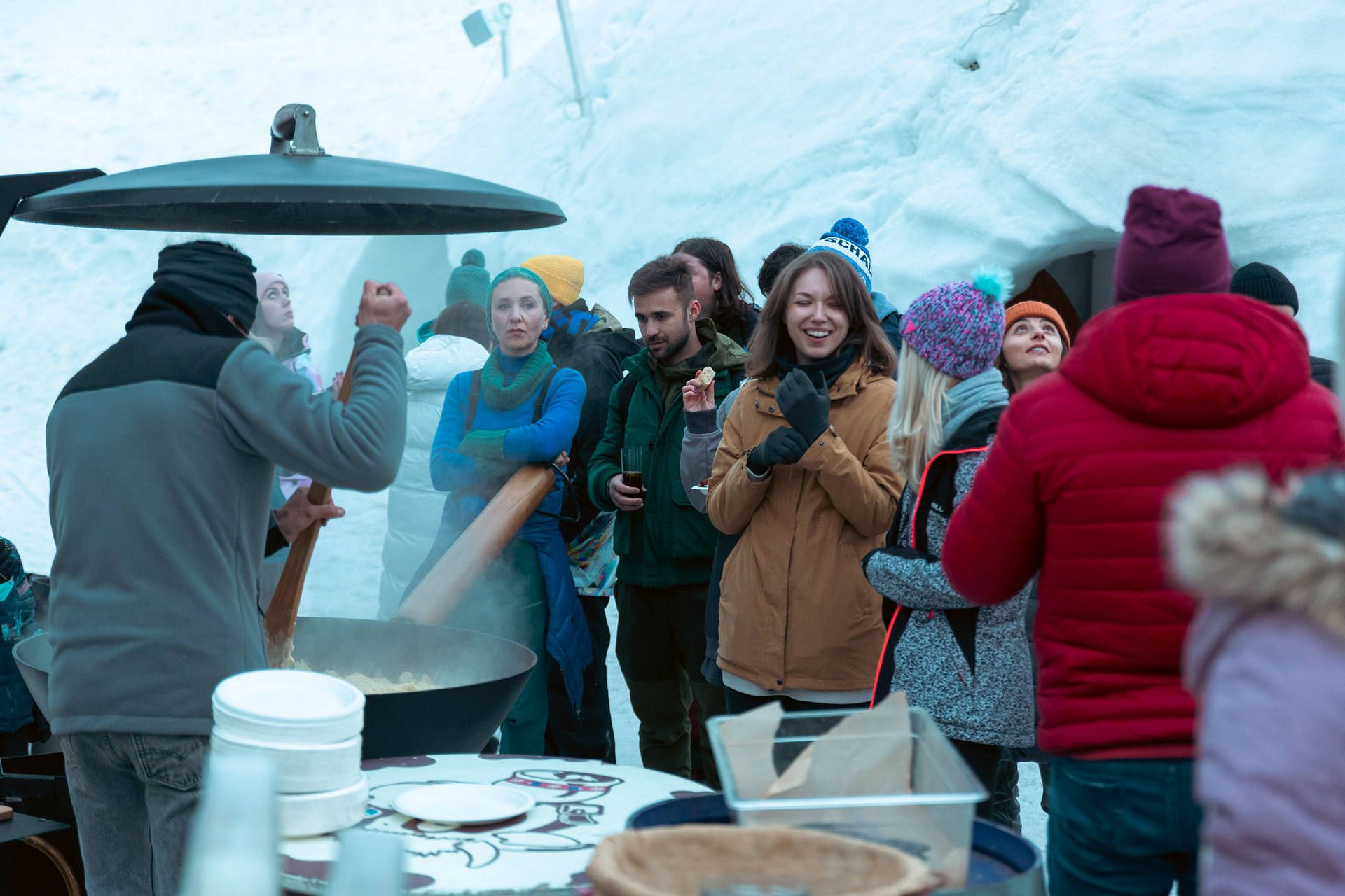 FJ7B6827 snow avoriaz | Fresh Planet | Fresh Planet Group of people in winter jackets gather around a large pot cooking outdoors inside an icy structure, sharing food and drinks.