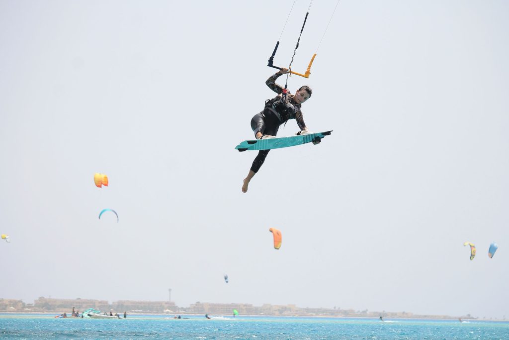 Kiteboarder mid-air performing a jump over blue ocean, turquoise board in hand, lines and bar overhead against a clear sky.