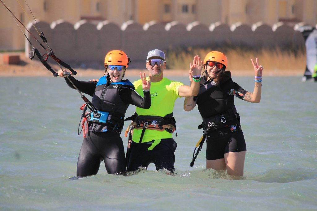 Three kiteboarders pose in waist‑deep water wearing orange helmets and wetsuits, smiling with raised hands after a session.