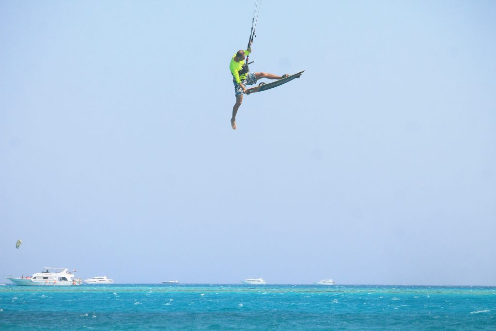 Kitesurfer in a neon shirt jumping high above turquoise water with boats in the background.