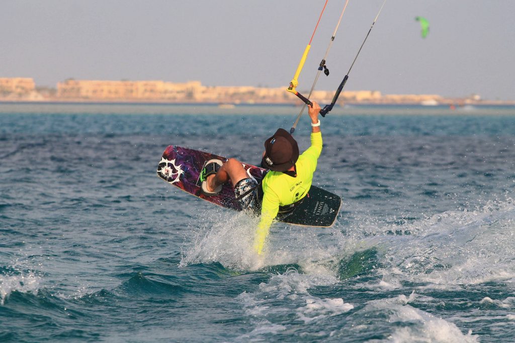 Kiteboarder in a neon lime shirt launches out of the water while holding the bar, with a distant shoreline in the background.