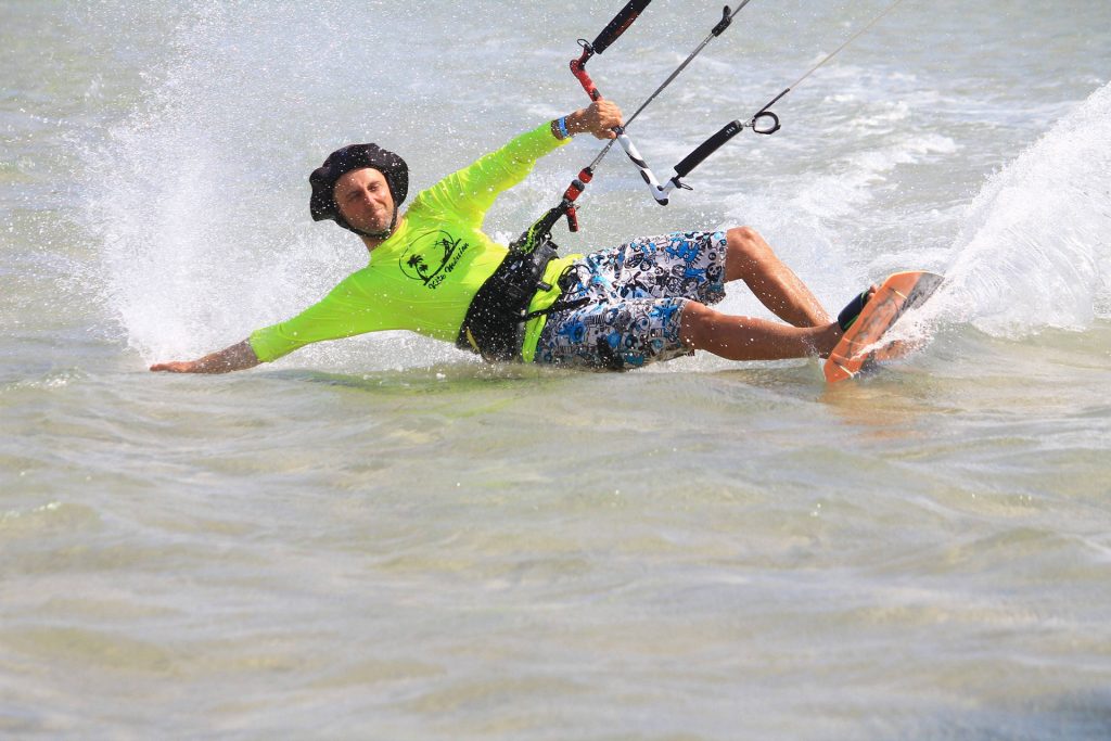 Kiteboarder in a neon lime shirt carving through shallow water, creating a large splash with the board tilted sideways.