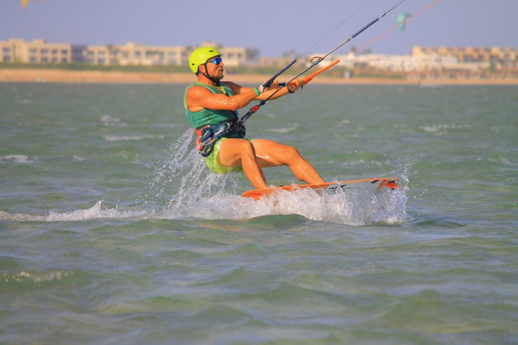 Kiteboarder in a lime helmet and green vest rides a board on choppy water, gripping orange and blue control bars as splashes fly.