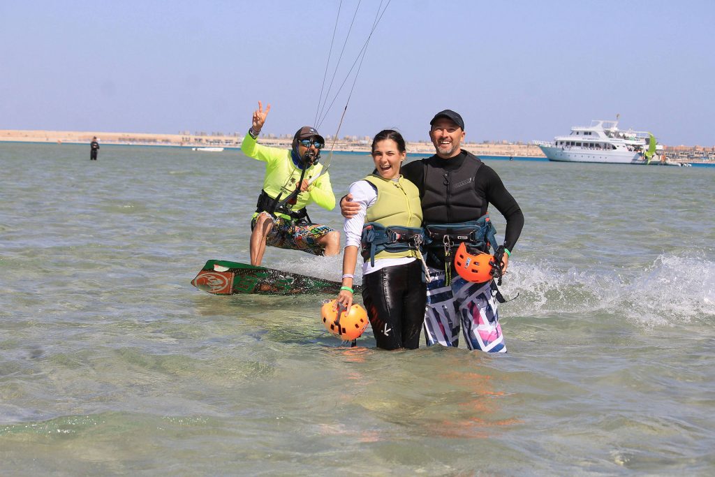 Group of three people kiteboarding in shallow water, two adults wearing harnesses and life vests, one making a peace sign in a neon shirt; orange helmets near them with a boat in the background.
