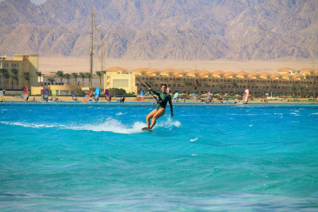 Kiteboarder riding turquoise sea near a resort beach with palm trees and rugged mountains in the background.