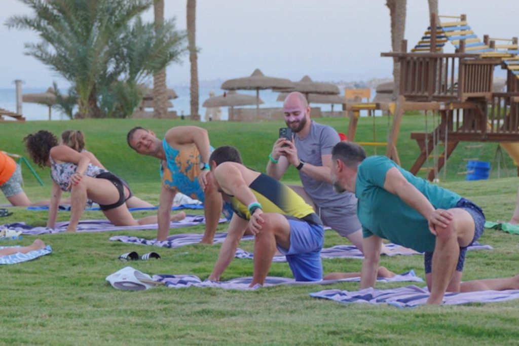 Group of people doing yoga on mats in a seaside park with palm trees and a playground in the background.