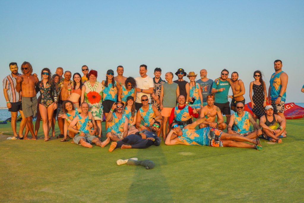 Large group of friends in colorful tropical outfits posing for a group photo on a sunny beach/grass area with a clear blue sky behind them.