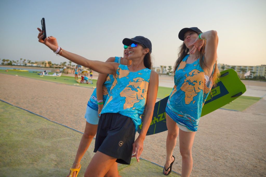 Two women in matching blue tropical outfits take a selfie on a sunny beachside boardwalk; one holds a smartphone aloft while the other poses with a wakeboard