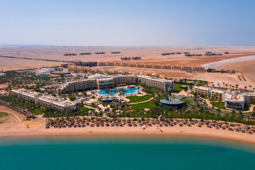 A large seaside resort with multiple buildings, a central pool, and palm-lined lawns along a turquoise beach.
