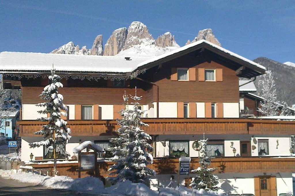 Snow-covered wooden chalet with a clear blue sky and snowy mountains in the background.