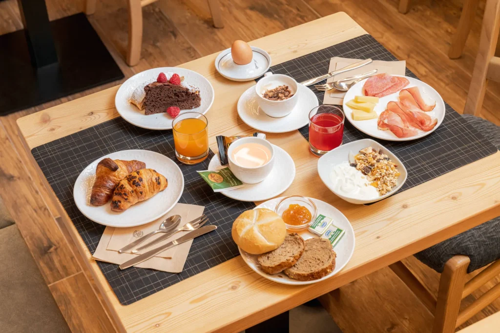 Breakfast spread on a wooden table: croissants, cake with raspberries, cheese, cold cuts, an egg cup, yogurt/granola, breads, and assorted drinks