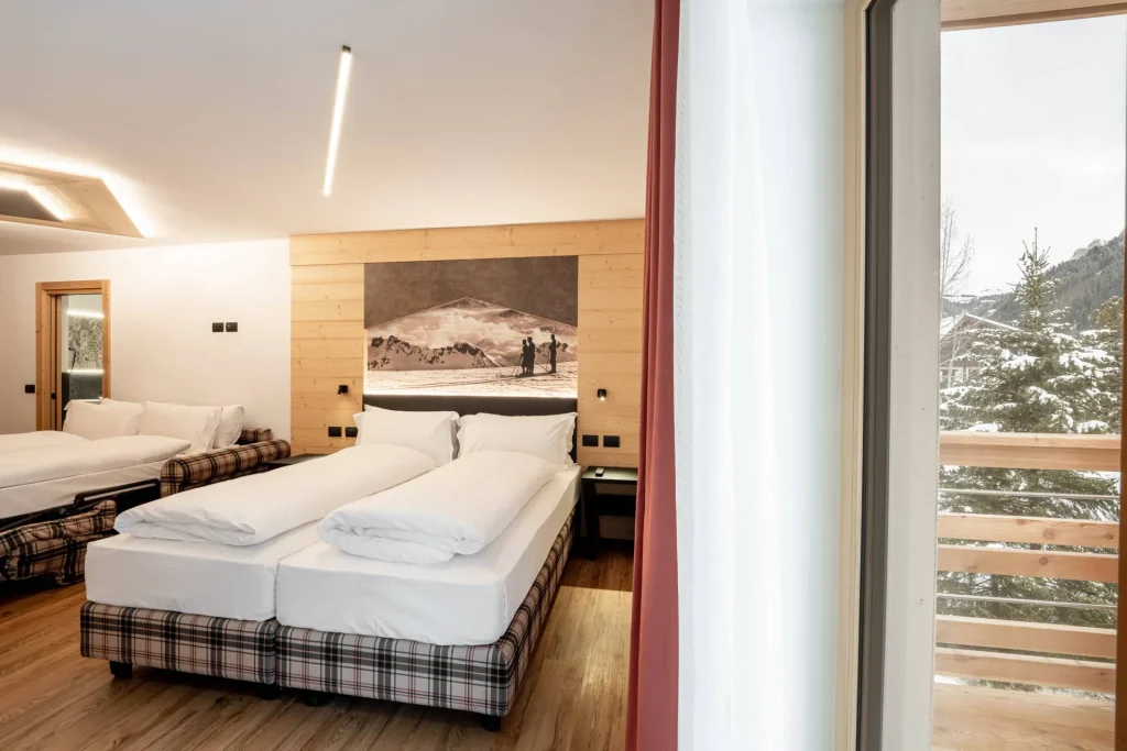 Hotel room with three white beds, plaid bases, wood panel wall, and a snowy mountain view through a large window.