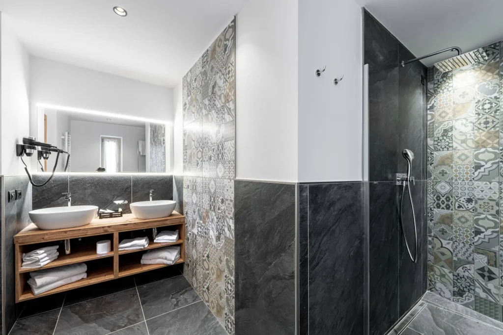 Modern bathroom with double vessel sinks on a wooden vanity, backlit mirror, and a tiled shower area with patterned ceramic tiles.