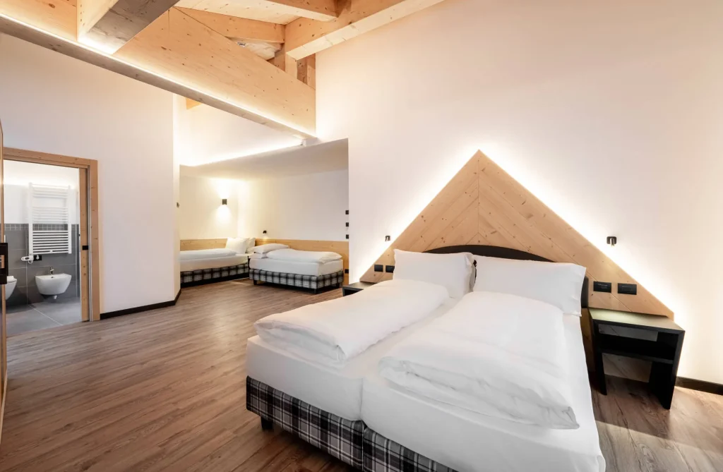Hotel room with three twin beds, light wood paneling, and exposed ceiling beams under warm lighting.