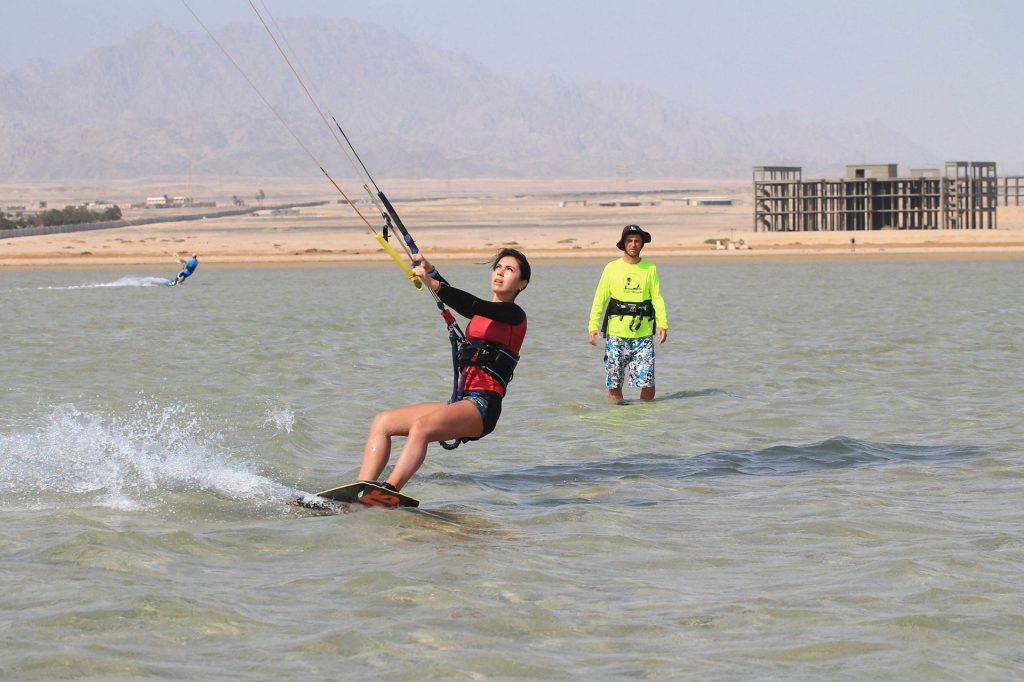 Woman kiteboarding in shallow water while a man watches nearby; desert shoreline and mountains in background.