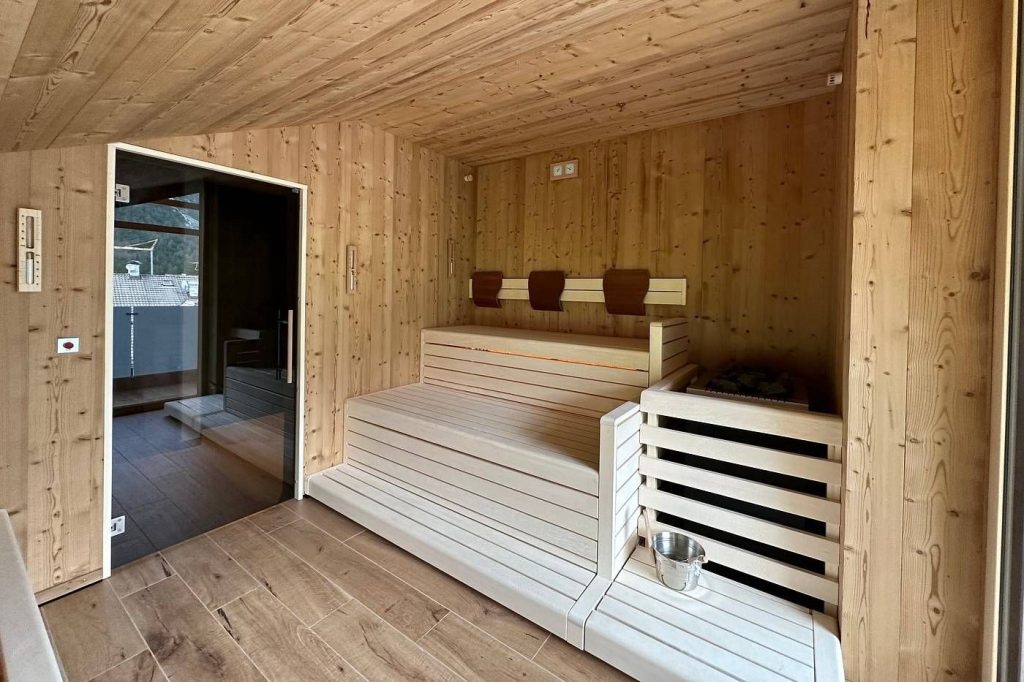 Wooden sauna interior with light benches, a heater on the right, and a metal bucket nearby.