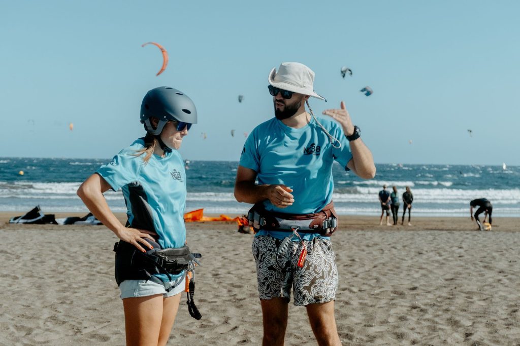 Two people on a windy beach in teal shirts with kiteboarding gear, helmeted and ready to surf, ocean in the background.