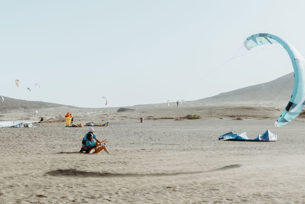 People on a sandy beach preparing paragliding gear; a large blue paraglider rests on the right with others in the distance under a clear sky.