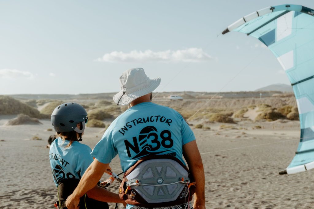 Two people in blue shirts stand on a sandy beach preparing for kitesurfing, one with a helmet and harness near a large kite.