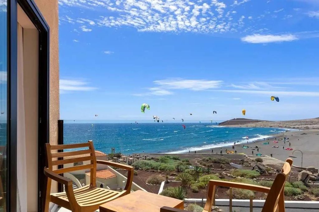 Ocean view from a balcony with wooden chairs; kite surfers ride the blue sea on a sunny day.
