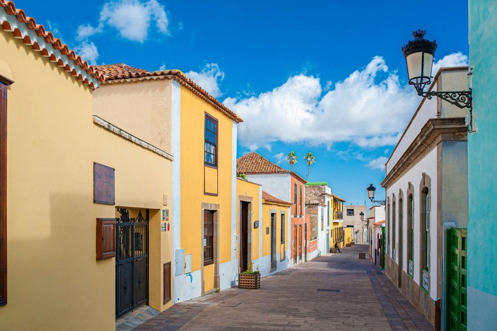 Colorful street in a coastal town: yellow and pastel houses with tiled roofs beneath a bright blue sky and vintage street lamps ahead of a palm-lined path to the distance.