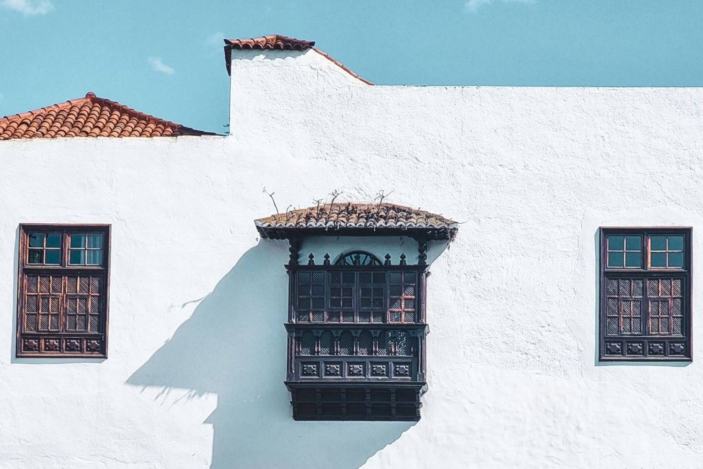 White stucco building facade with two dark wooden-framed windows on sides and a central ornate black balcony under a tiled roof, casting a shadow on the wall