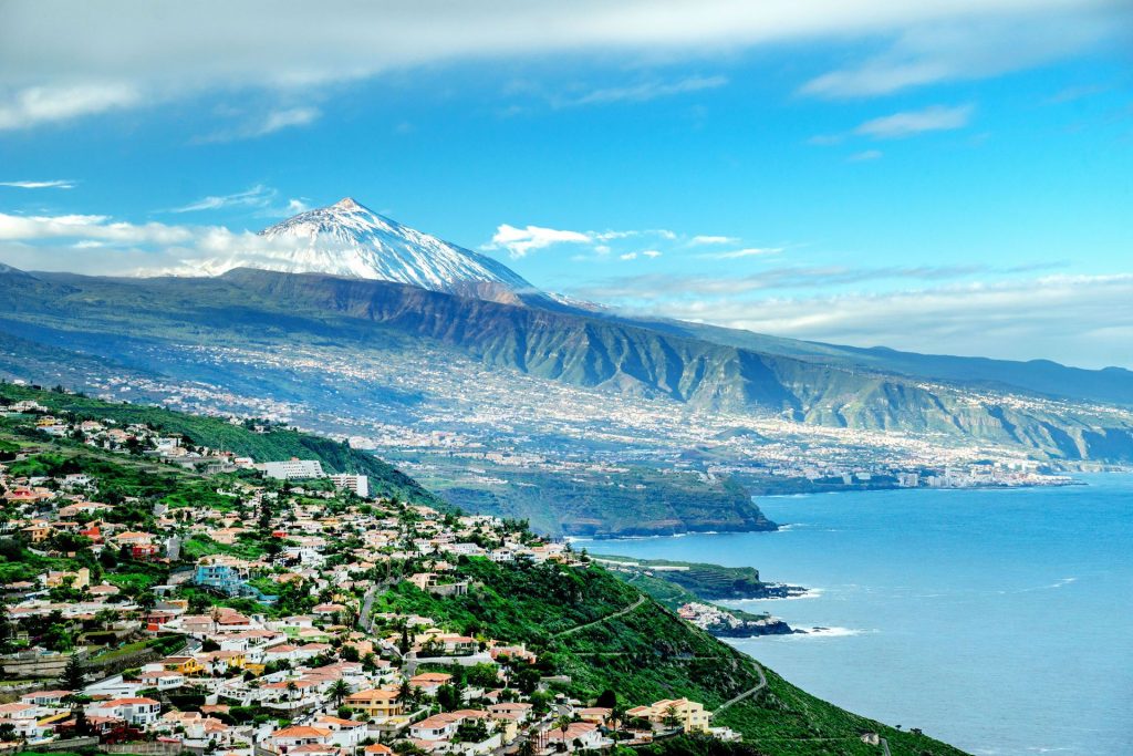 Coastal hillside town with colorful houses cascading toward the sea and a snow-capped mountain in the distance under a blue sky