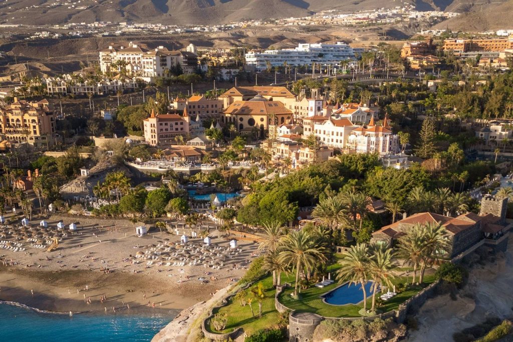 Aerial view of a seaside resort with peach-colored buildings, palm trees, and a curved pool beside a sandy beach.