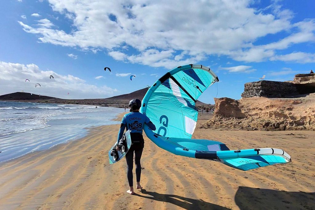 Kitesurfer in a blue wetsuit walks along a sandy beach carrying a large turquoise kite with the ocean on the left and rocky cliffs in the background.