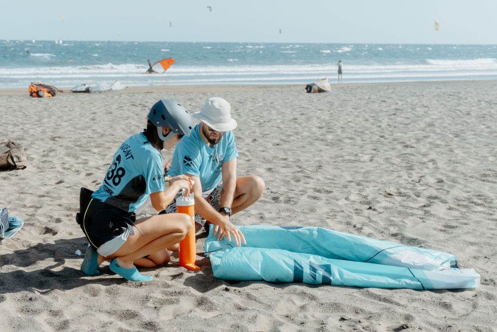 Two people in blue shirts sit on the sand, assembling windsurfing gear beside a folded blue sail.