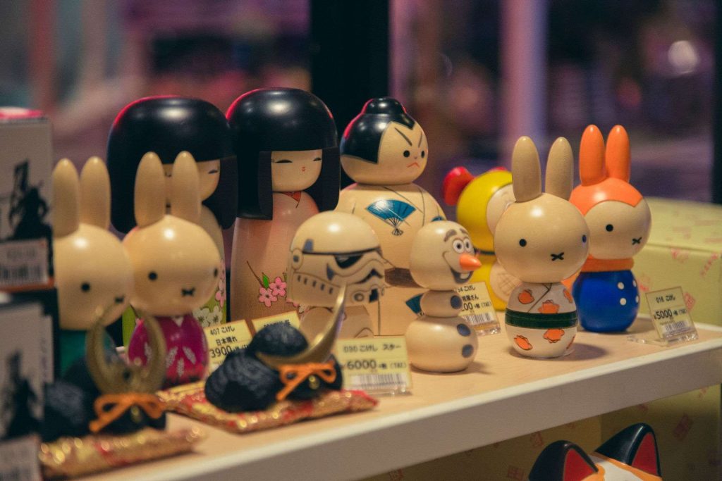 Row of colorful wooden toy dolls with painted faces on a shop shelf, price tags in Japanese visible in front of them.
