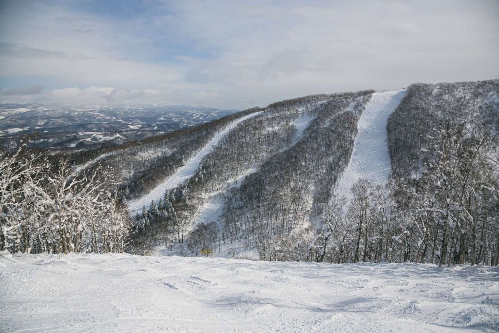 Snow-covered mountain slope with a groomed ski trail winding through bare trees, under a cloudy blue sky and distant hills far in the background.