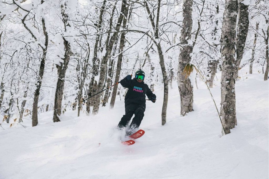 Skier in a helmet and bright green goggles skiing through a snow-covered forest between snow-dusted trees.