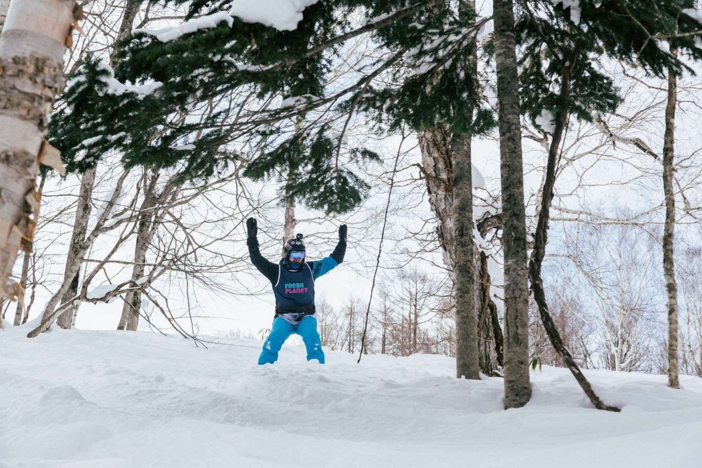 A skier with arms raised snowplows down a snowy slope between bare trees in a forested winter scene.