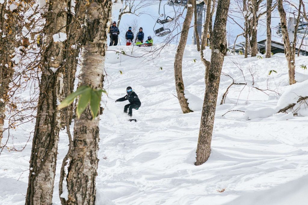 Snowboarder in black gear carving down a snowy slope through trees, while a small group watches from the crest and a chairlift looms above behind them.