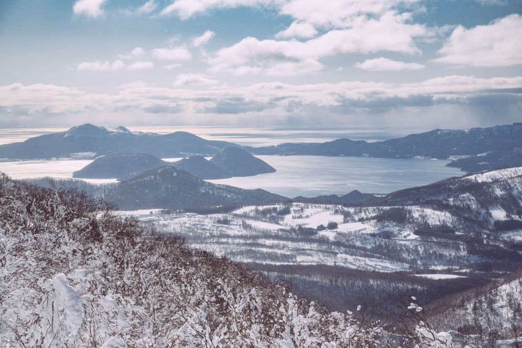 Snowy mountains surround a frozen lake, with snow-dusted shrubs in the foreground and a pale blue, cloud-filled sky above.