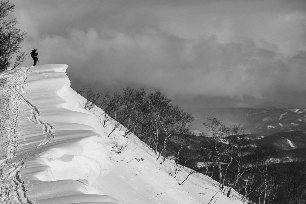 Person standing on a snowy cliff edge with trekking poles, looking over a mountainous landscape in winter (black and white).