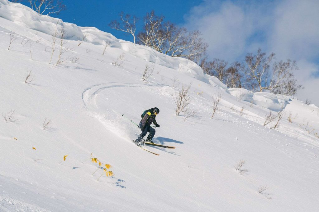 Skier in a black and yellow jacket carving down a snow-covered slope under a blue sky inside a wintry landscape.