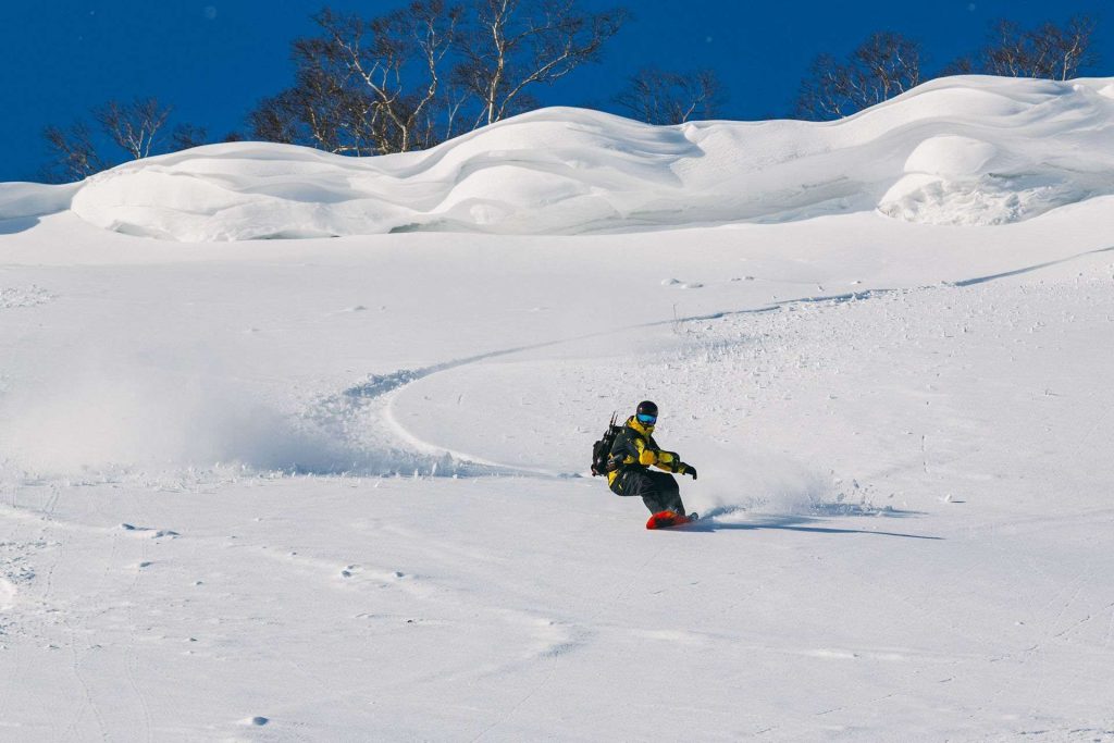 Snowboarder in a yellow jacket carving down a snowy slope with a blue sky and bare trees in the background.