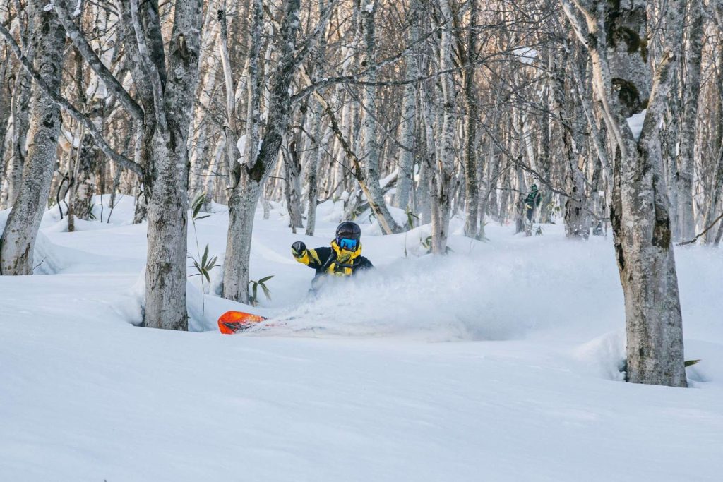 Snowboarder in a bright yellow jacket and blue goggles carving through fresh powder in a snow-covered forest of bare trees.