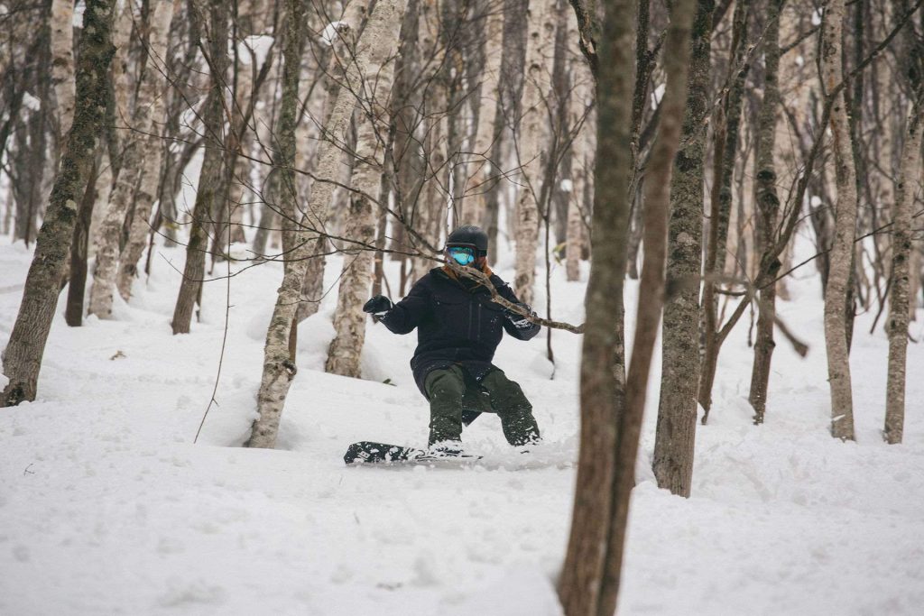 Snowboarder in a black jacket and green pants carving through a snowy forest among slender trees, wearing a helmet and blue goggles.